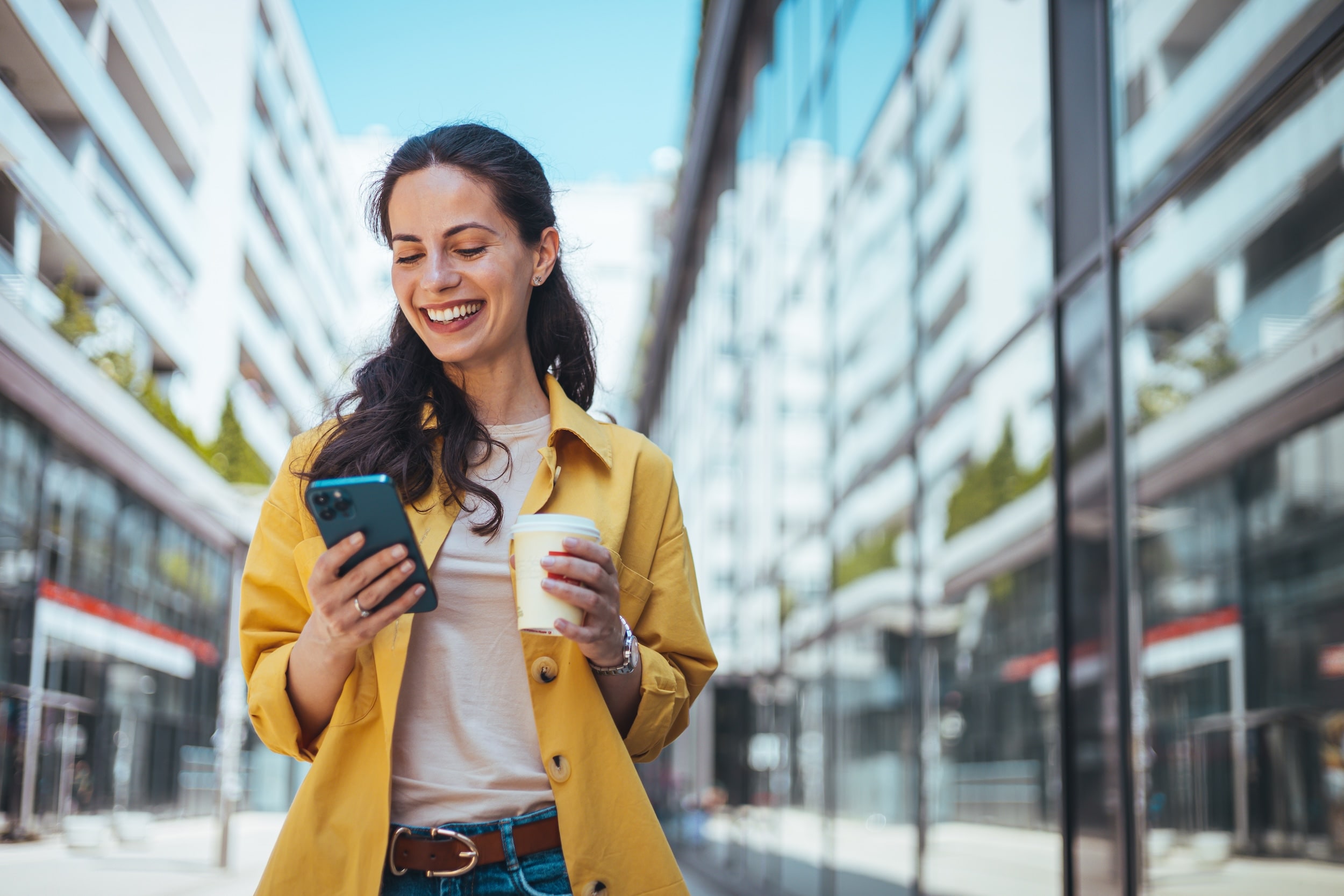 Woman in yellow jacket looking at her phone with coffee in hand