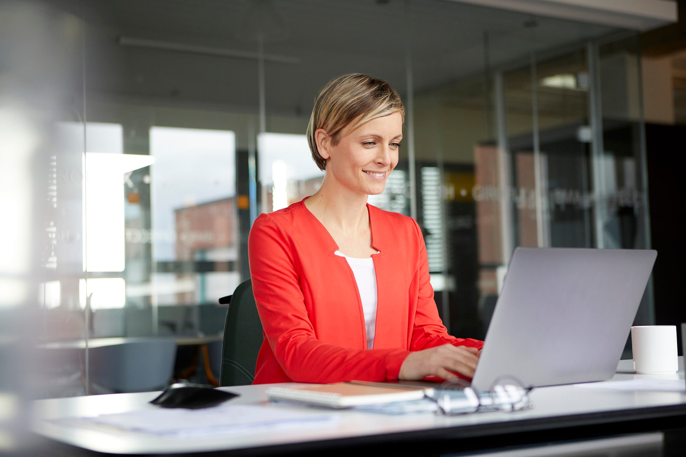 Lady with short blonde hair working in an office on her lap top, wearing a red blouse