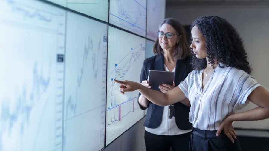 One woman pointing at giant screen and another woman with clipboard next to her